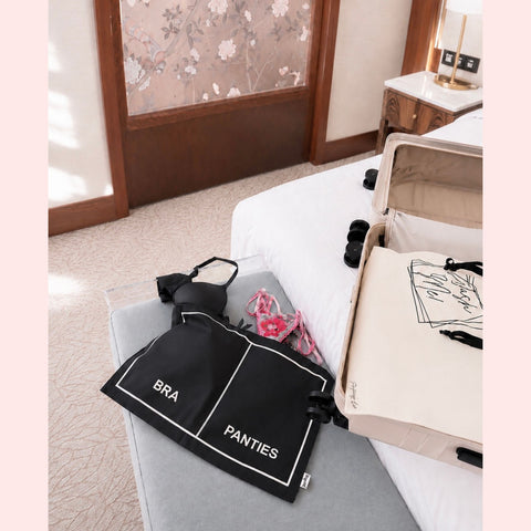 Black bags labeled 'BRA' and 'PANTIES' on a gray surface with an open suitcase in the background.