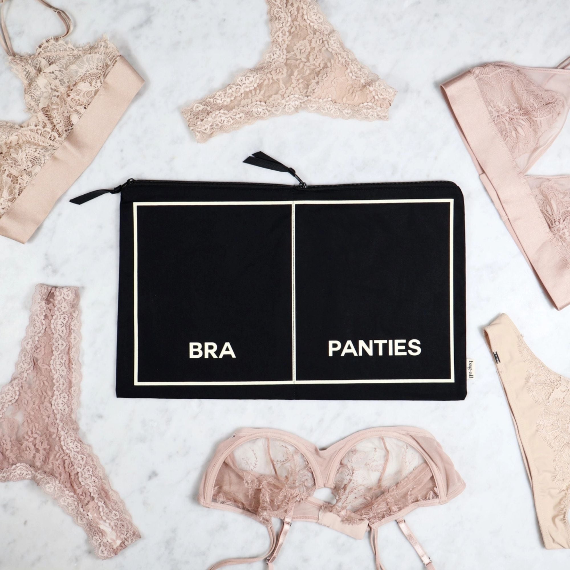 Black zippered pouch labeled 'BRA' and 'PANTIES' surrounded by lace lingerie on a light background
