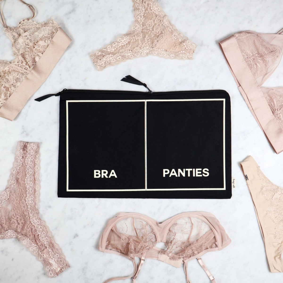 Black zippered pouch labeled 'BRA' and 'PANTIES' surrounded by lace lingerie on a light background